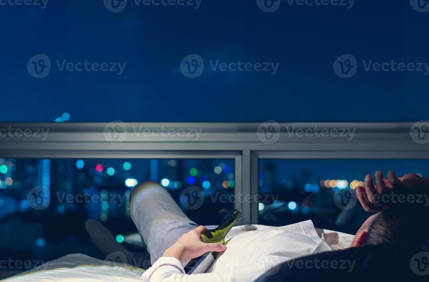 The stressed man lying on bed with a bottle of red wine and background of city bokeh lights at night. photo