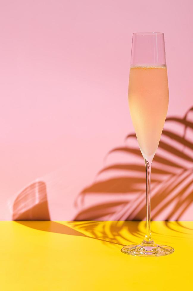 A glass of cold Champagne with vapour that has coconut leaf shadow from sun light on pink and yellow background. Summer concept. photo