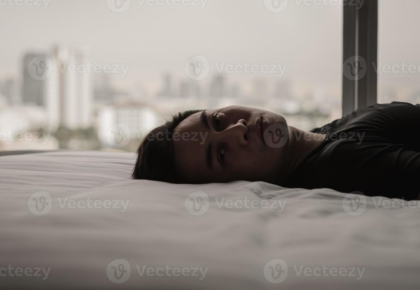 Asian man feels sad alone on bed with city background. Stay home, depression and loneliness concept. photo