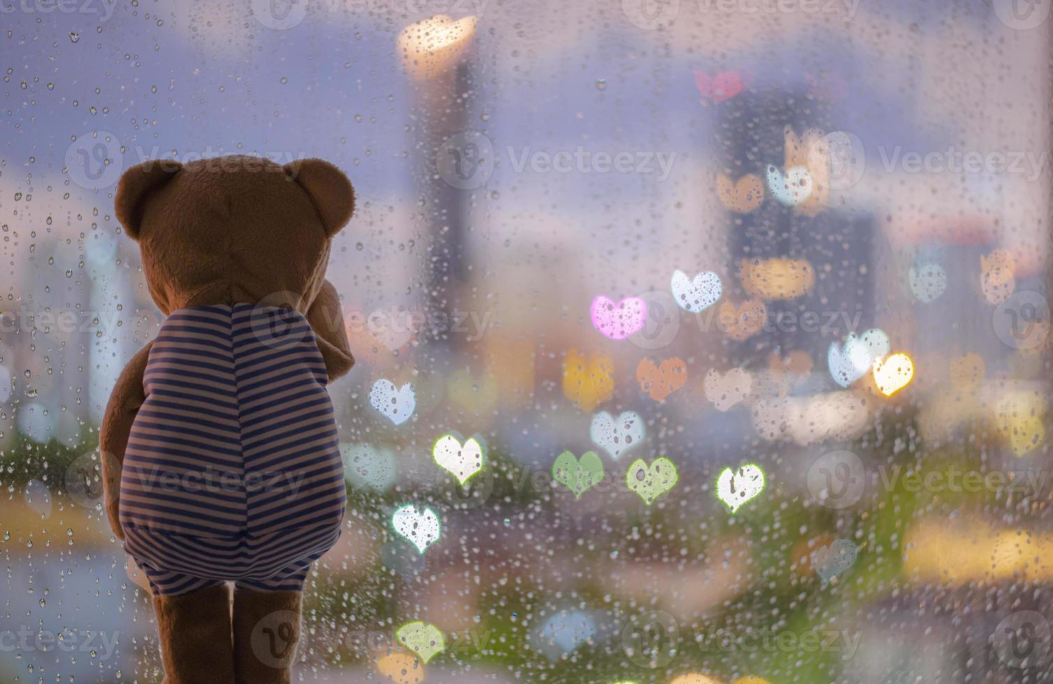 Teddy bear crying alone at window when raining with colorful love shape bokeh lights. photo