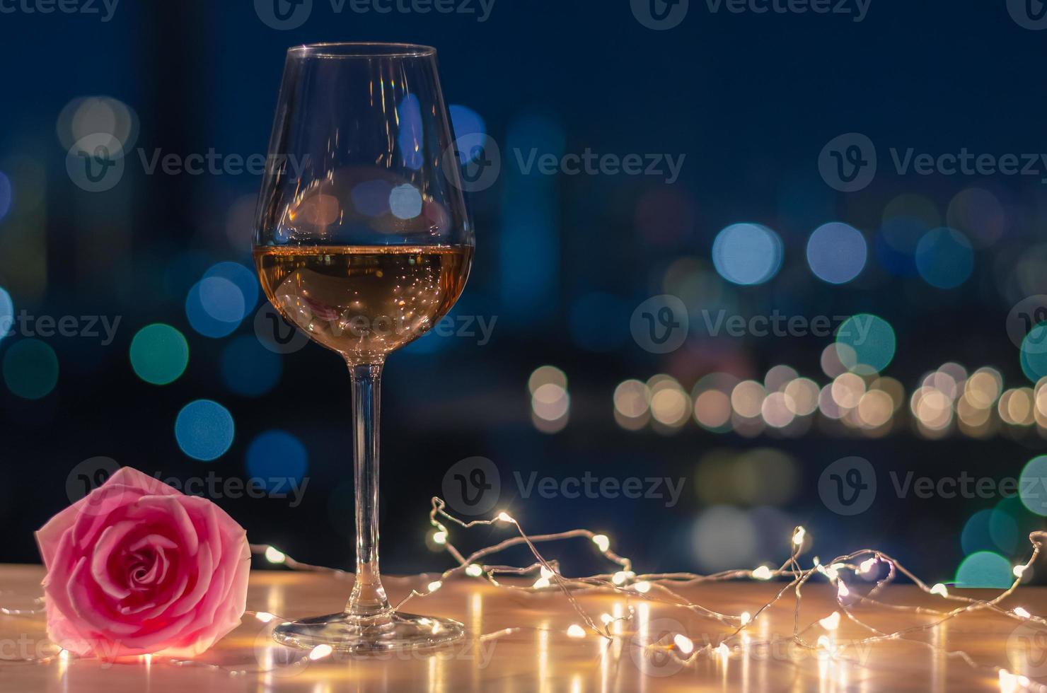 una copa de vino rosado con flor de rosa en la mesa y un colorido fondo claro de la ciudad. foto