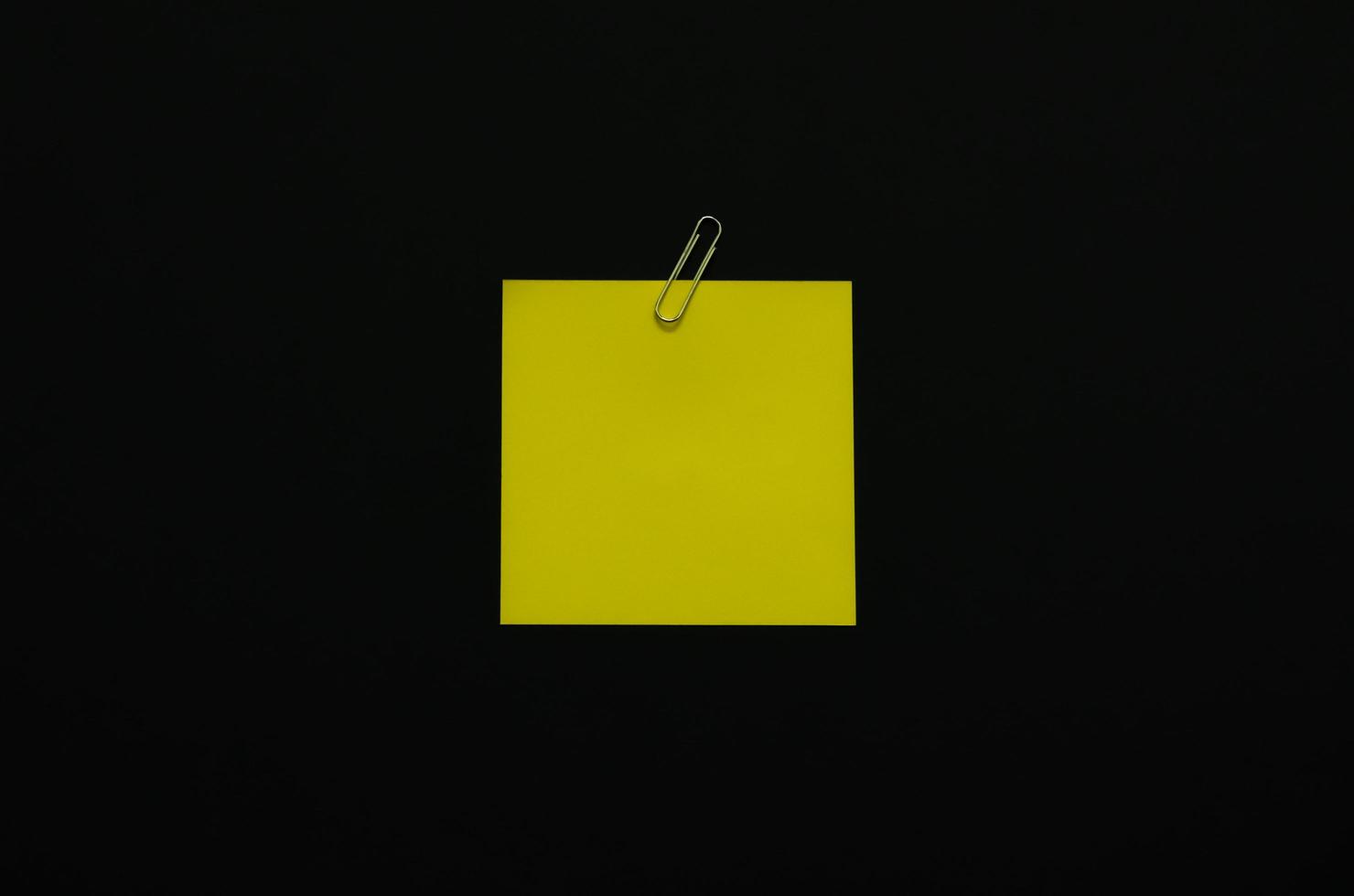 Yellow blank note paper with paper clip on dark background. Minimalist flat lay black concept. photo