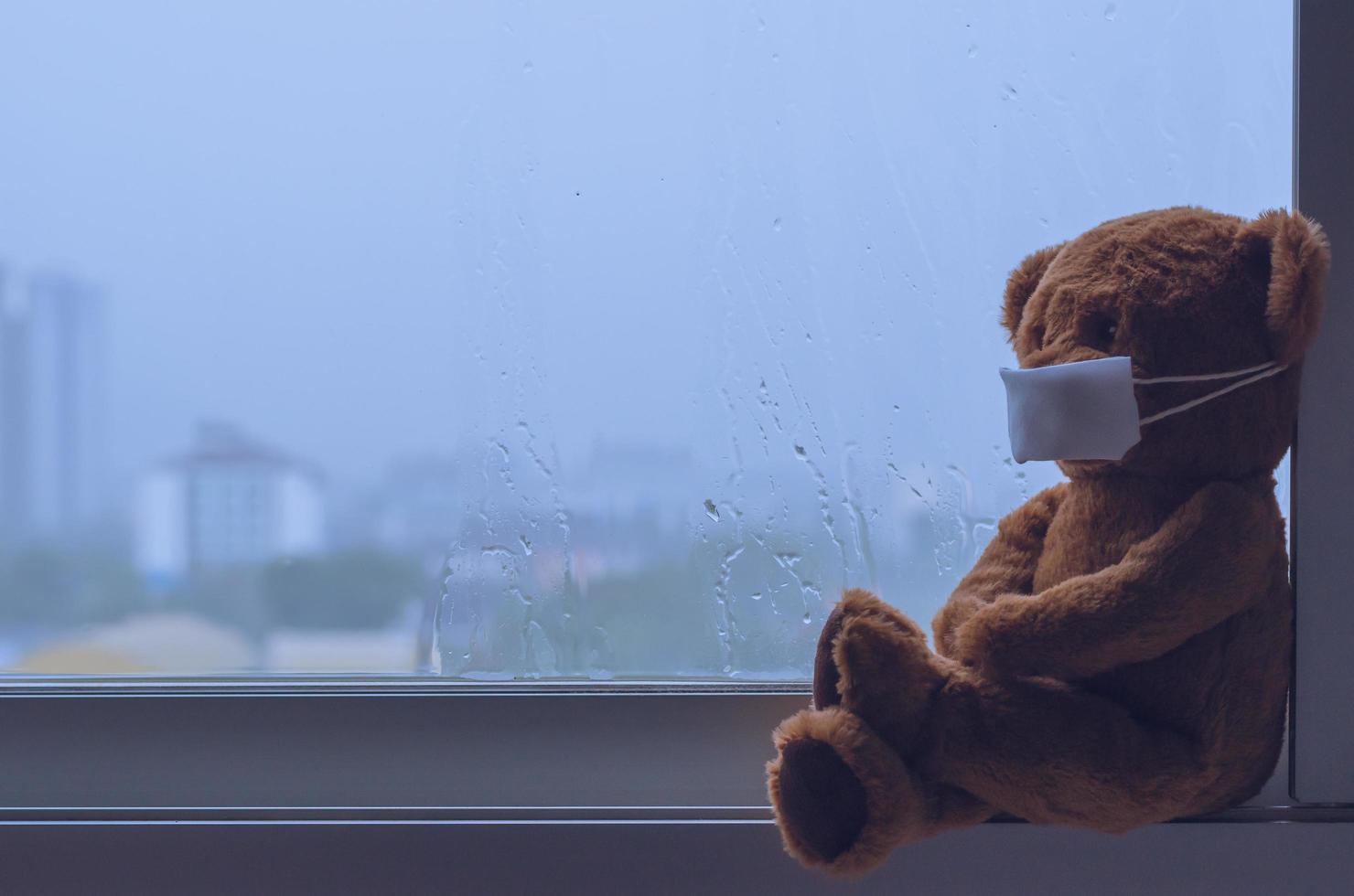 Teddy bear wearing mask sitting at window while raining in monsoon season. photo
