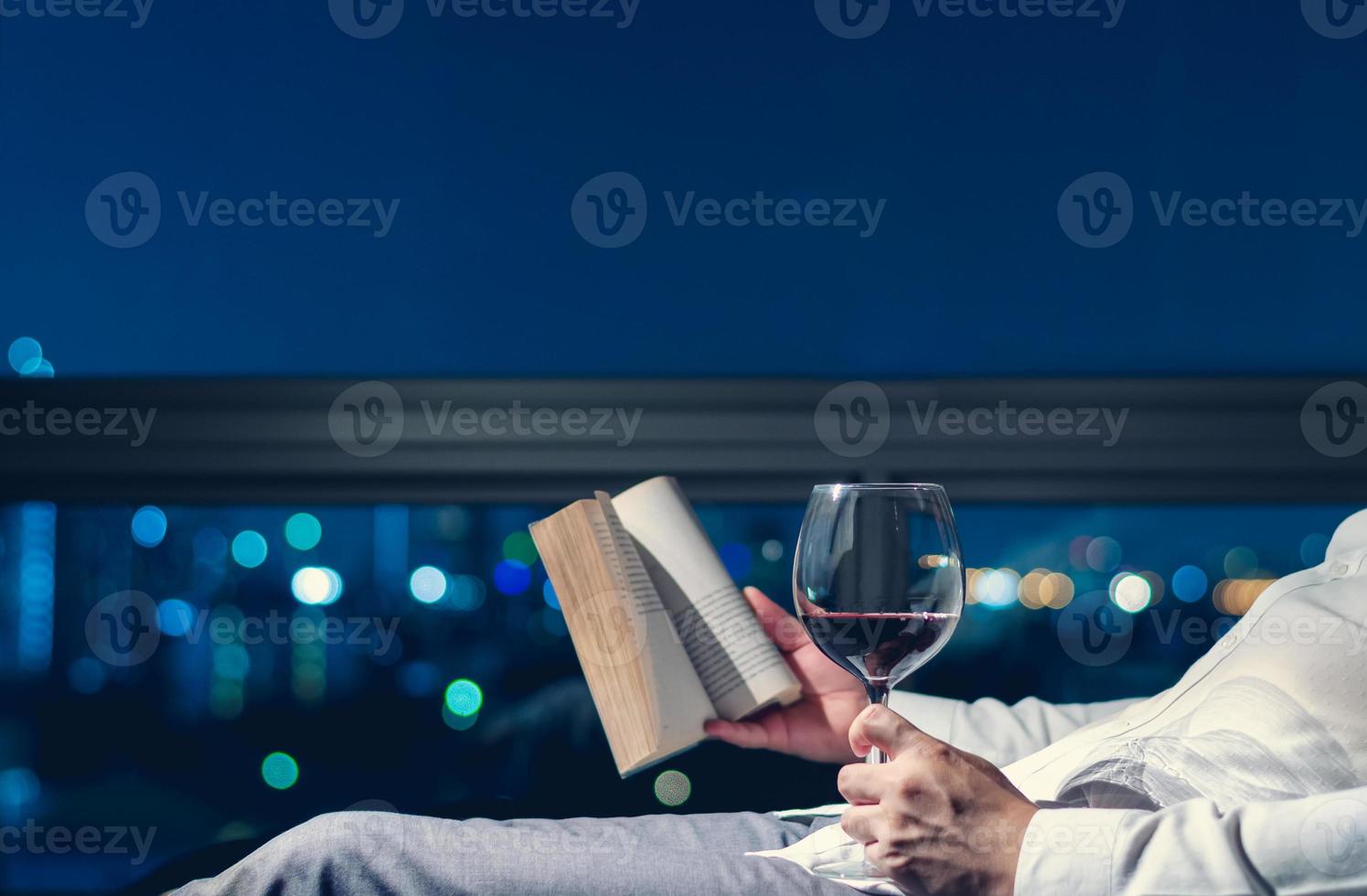 The man relaxing in the room reading book with a glass of red wine and background of city bokeh lights. photo