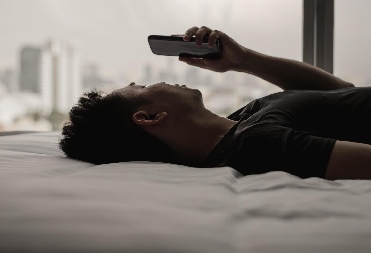 el hombre asiático se siente triste en la cama mirando y concentrándose en el teléfono inteligente. adicción a las redes sociales de internet y concepto de salud mental. foto