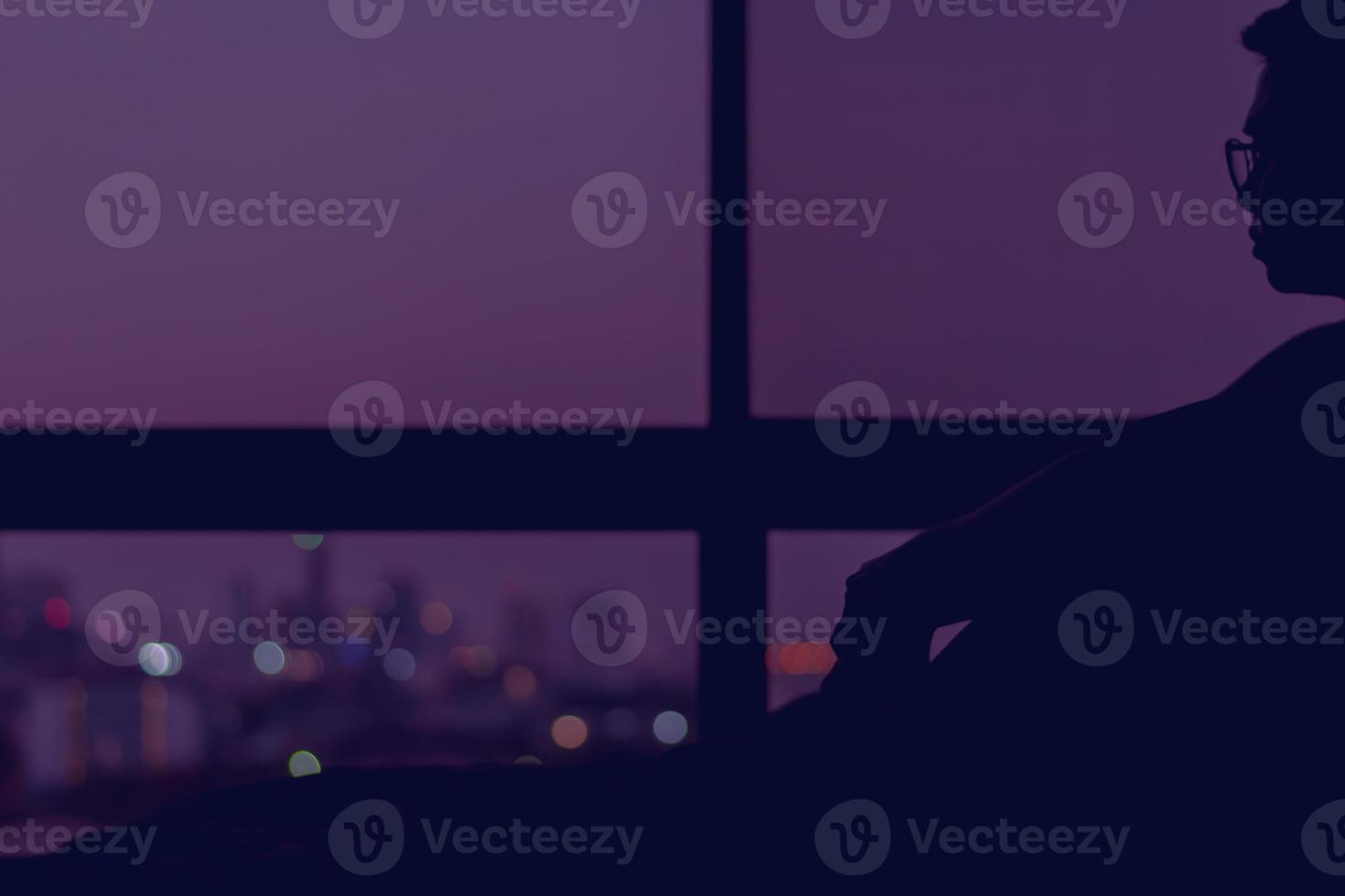 A man sitting on his bed alone at night looking out at colorful city bokeh lights. photo