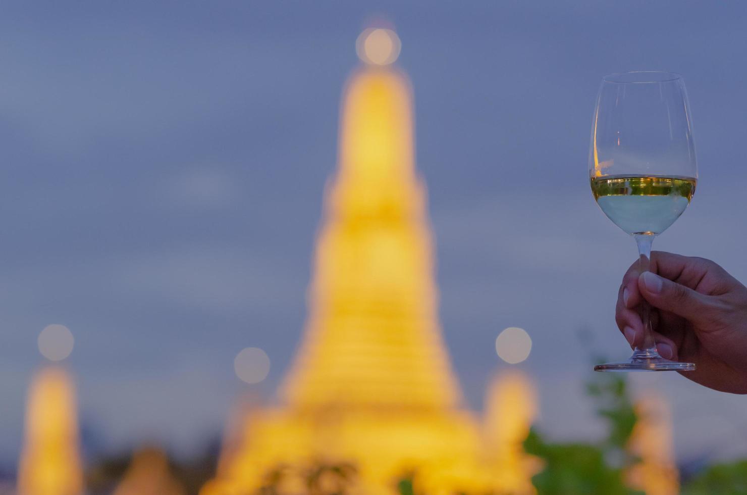 Hand toasting a glass of white wine with blurred background of temple at night. photo