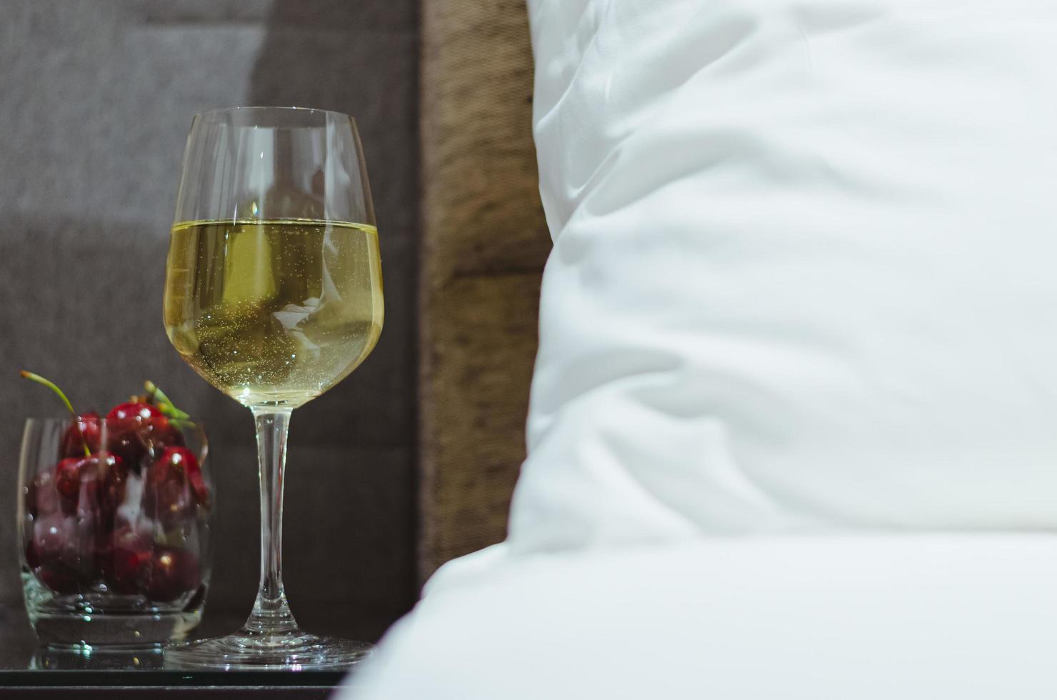 A glass of white wine with cherry fruits put on table besides bed with white bedsheet. photo