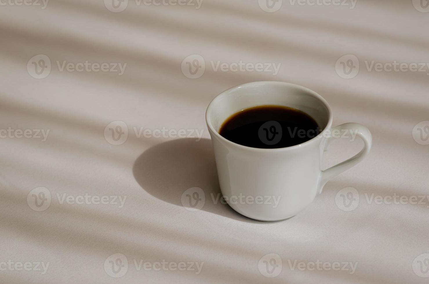 A cup of black coffee puts on white bed sheet with shadow of curtain in the morning. Relaxing at home concept. photo