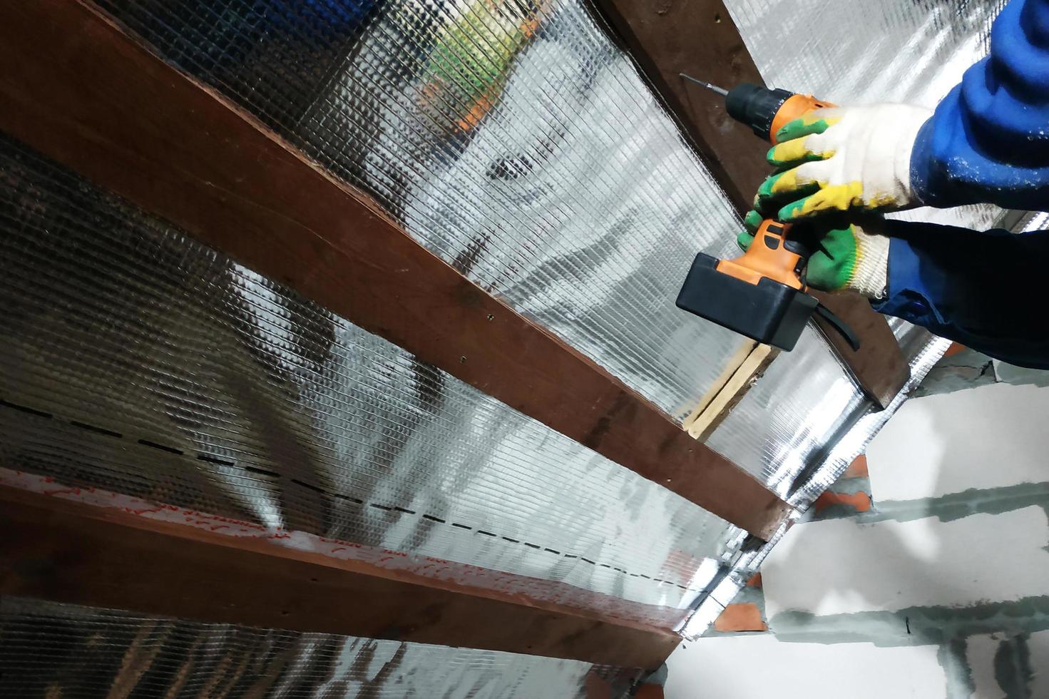 The wizard uses a cordless drill and screw the screws into boards that hold foil film and mineral wool to heal the roof of the house photo