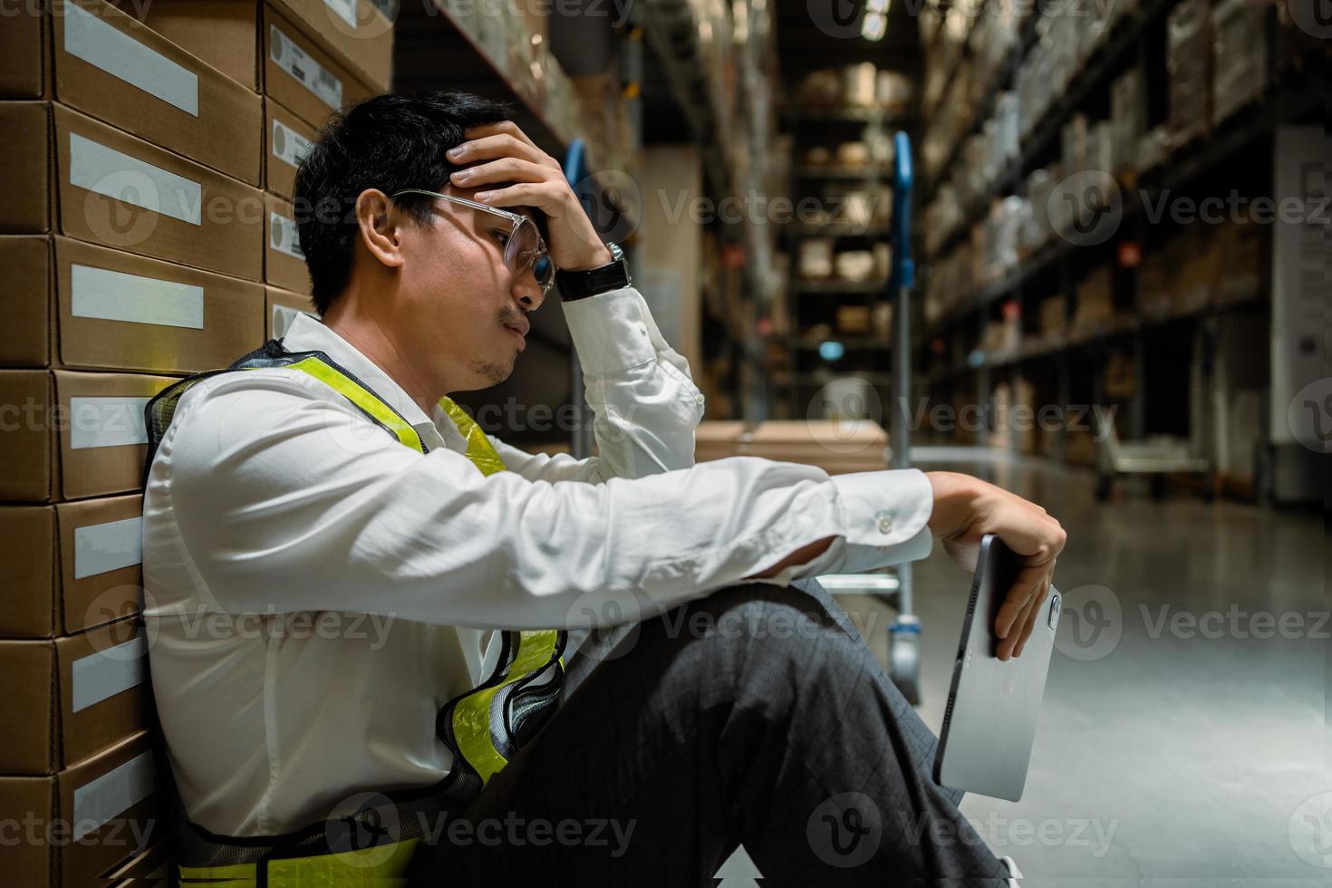 An workers anxious leans against a box in a warehouse.. Hold the tablet and think about work problems and hold your head in the warehouse. Warehousing and logistics concepts photo