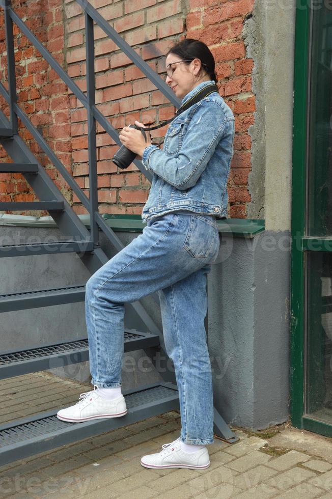 A young woman photographer in a blue denim jacket and glasses stands against the background of a brick wall, with a camera in her hands and looks through photos. Girl with a camera. photo