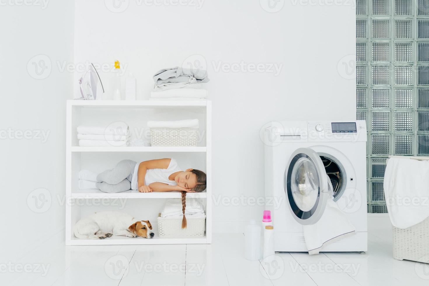 Cozy washing room with washer, sleeping girl with dog on shelf, bottles of liquid powder on floor, basket full of dirty laundry. Child has rest after helping mum to wash clothes. Domestic atmosphere photo