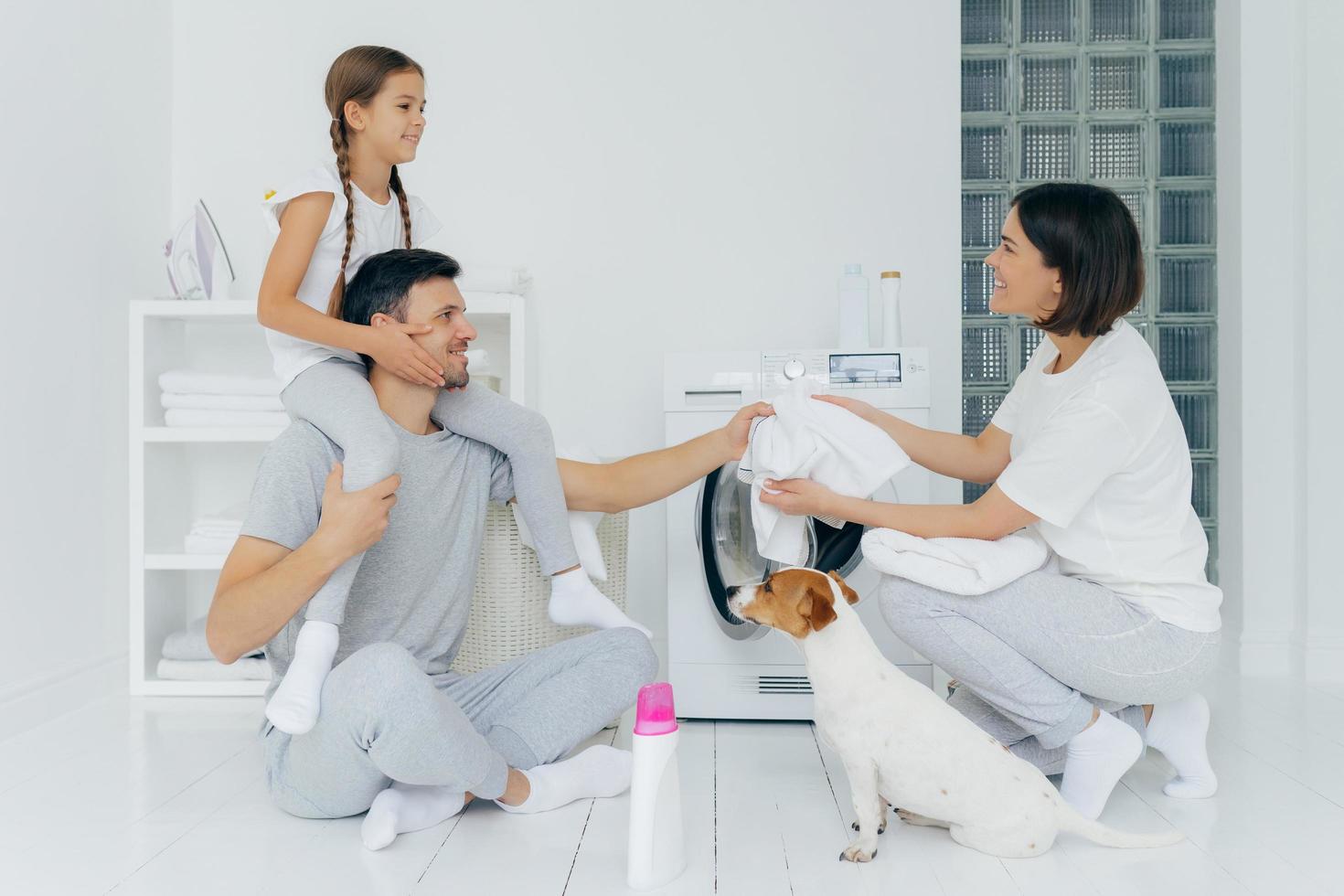 Horizontal shot of little girl has fun together with parents, rides on fathers back, pose together near washing machine in laundry room, dog near, load laundry into washer, busy with domestic work photo