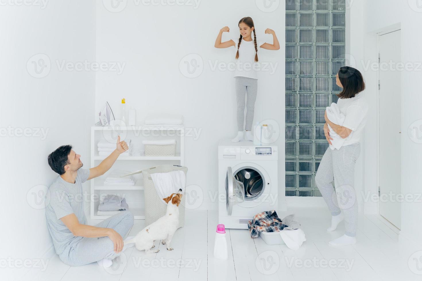 Image of glad small child raises arms, shows biceps and stength, father shows like sign with thumb up, stand in washing room with pile of clothes in basin near washer, detergent. Well done work photo