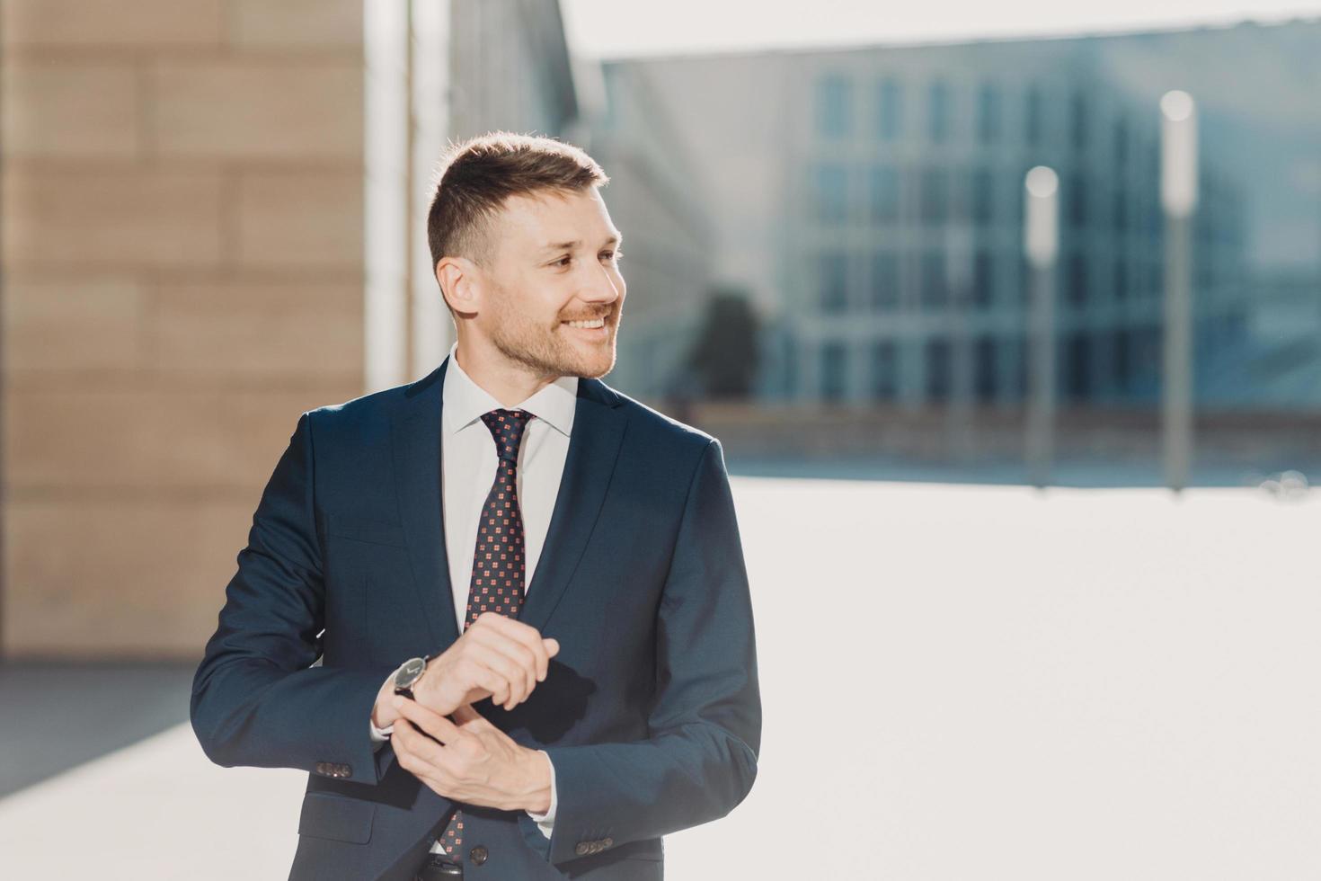 People and business concept. Glad elegant young businessman with stubble, wears formal clothes and watch, waits for someone, poses outdoor near office. People, career and leadership concept. photo