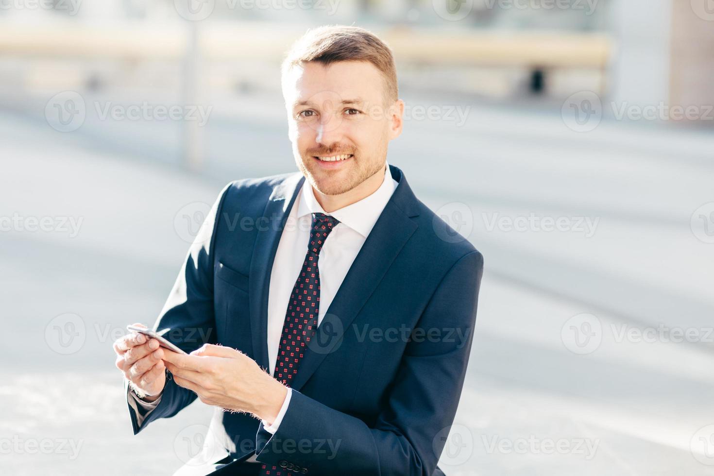 imagen al aire libre de un exitoso empresario masculino, usa traje negro formal, camisa blanca con corbata, sostiene un teléfono inteligente y revisa la notificación en la calle, envía un mensaje de texto o revisa el buzón de correo electrónico en línea foto