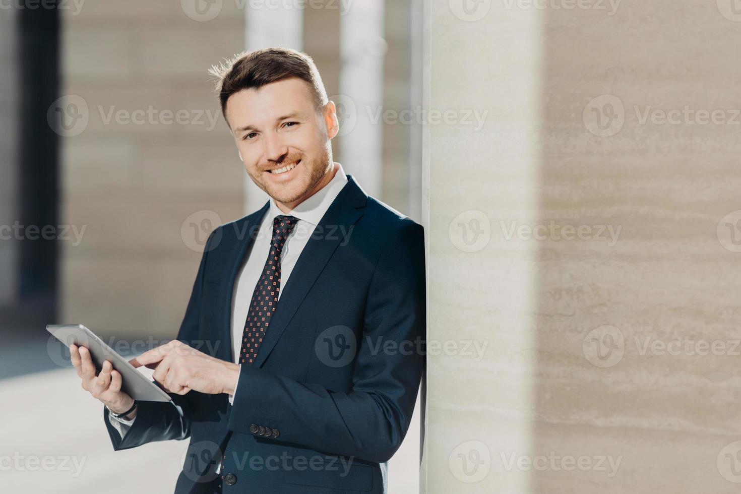 Experienced male economist in formal clothes works on digital tablet, has work break, stands in office interior with copy space. Successful entrepreneur prepares business report on modern device photo
