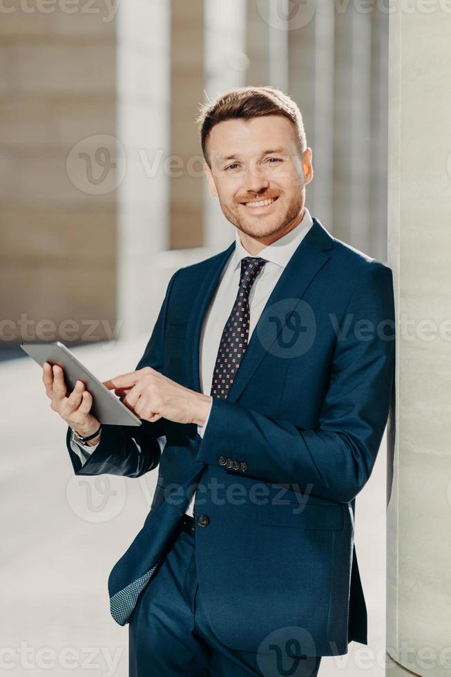 Vertical shot of business person uses digital tablet for online communication and work, uses free internet at office, has positive expression, dressed in formal black suit. People and occupation photo