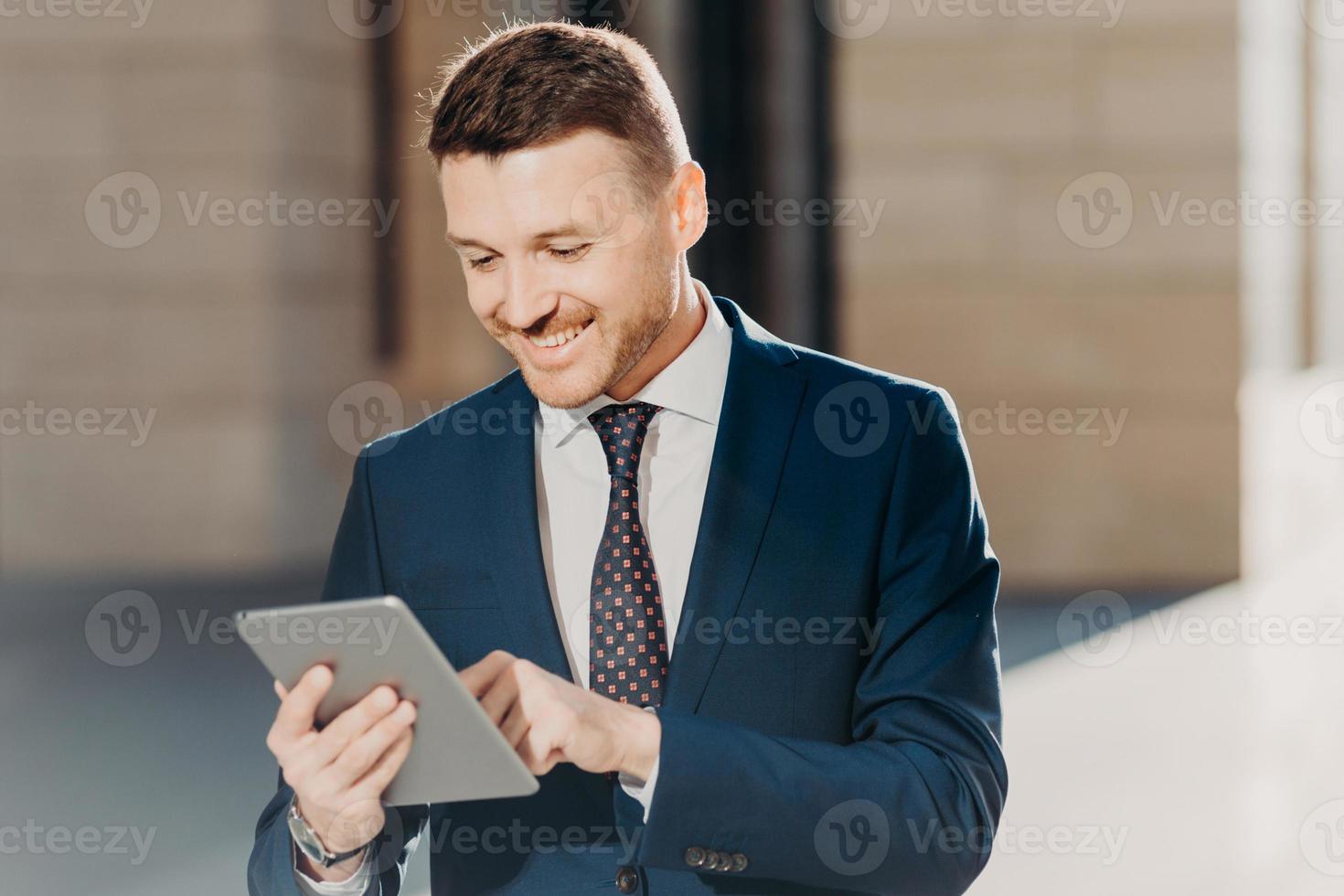 Confident handsome male manager checks email on tablet computer, updates database on touch pad, wears elegant formal clothing, connected to wireless internet, poses outdoor. Business concept photo