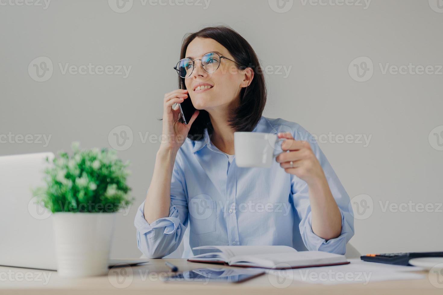Gorgeous pleased businesswoman has cell phone conversation while has coffe break after working on notebook, shares her success with close person, isolated over grey background. Occupation concept photo