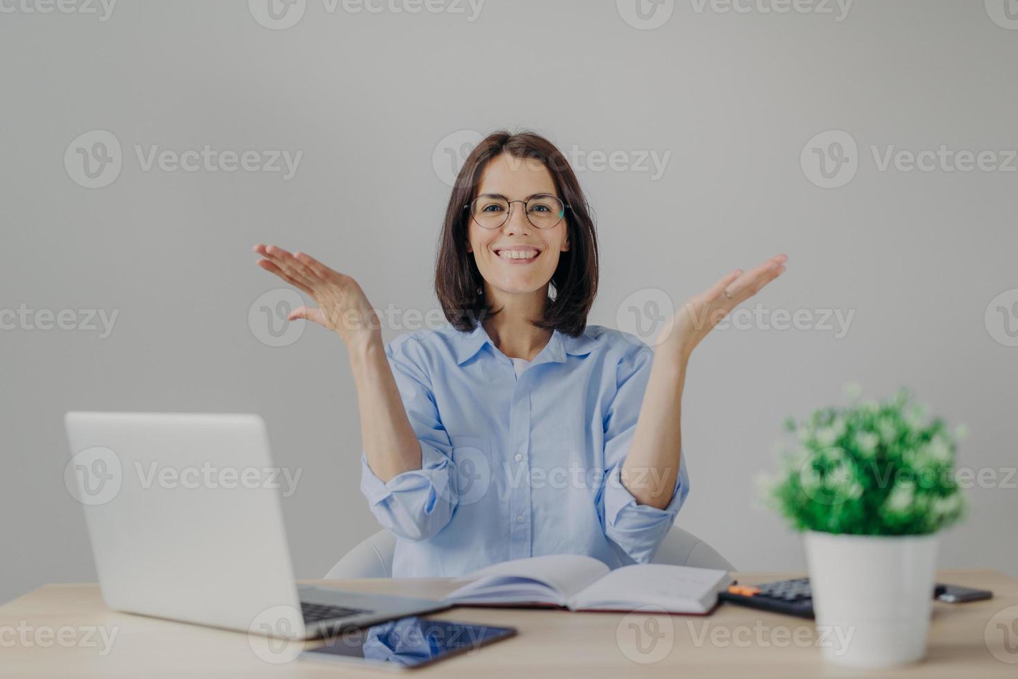 Happy overjoyed female achieves success in her work, going to present high results, raises hands joyfully, sits at working place with laptop computer, diary, calculator, isolated on grey background photo