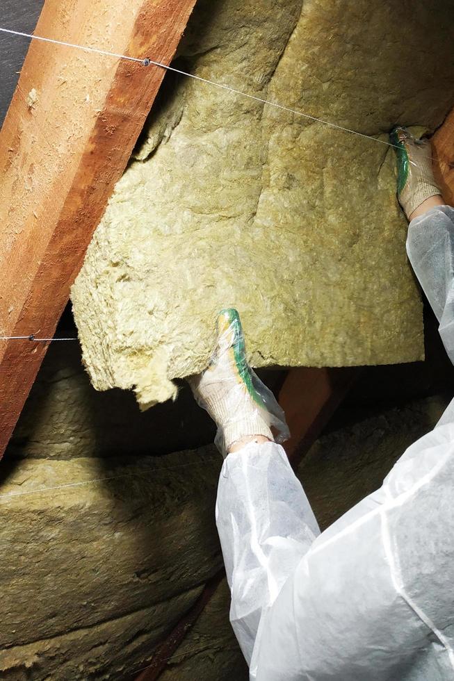 A man in a protective overalls puts mineral wool between the beam of the roof of the house for his warming from the cold photo