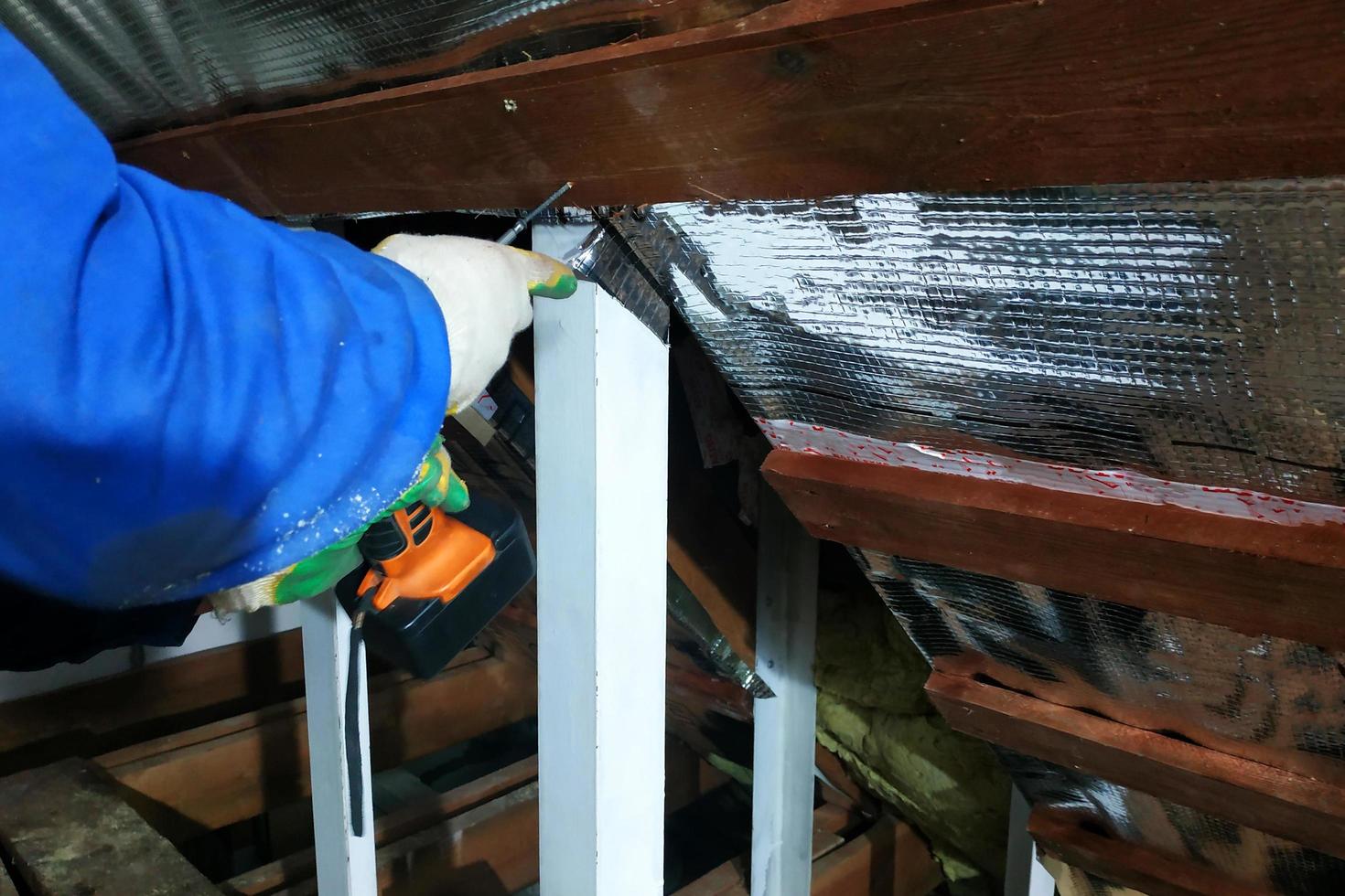 The wizard uses a cordless drill and screw the screws into boards that hold foil film and mineral wool to heal the roof of the house photo