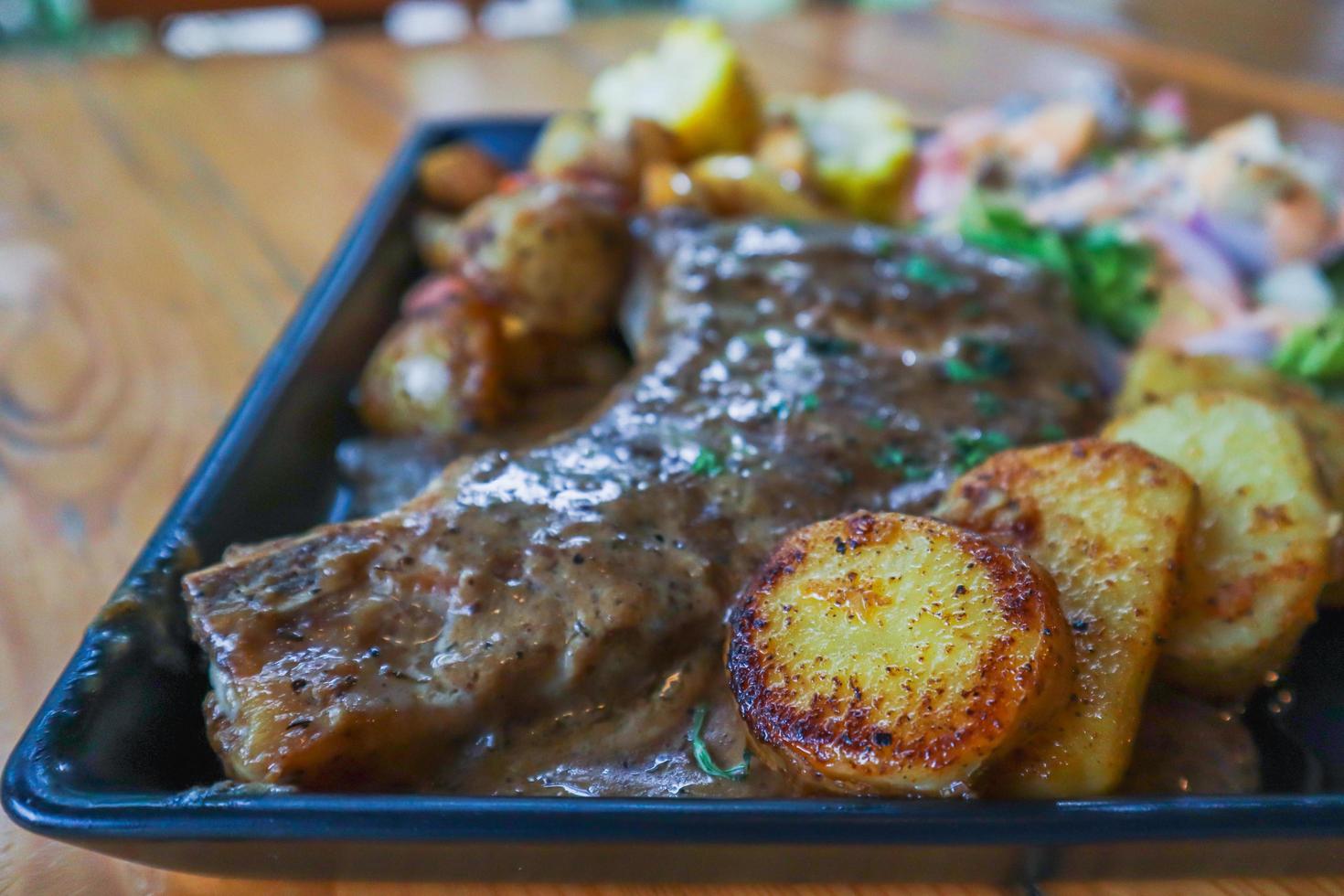 Grilled beef steak with gravy is served with a fruit and vegetable salad inside a black ceramic plate on the dining room table to prepare the steak for a festive dinner. photo