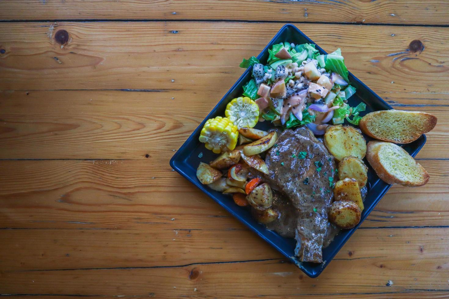 Grilled beef steak with gravy is served with a fruit and vegetable salad inside a black ceramic plate on the dining room table to prepare the steak for a festive dinner. photo
