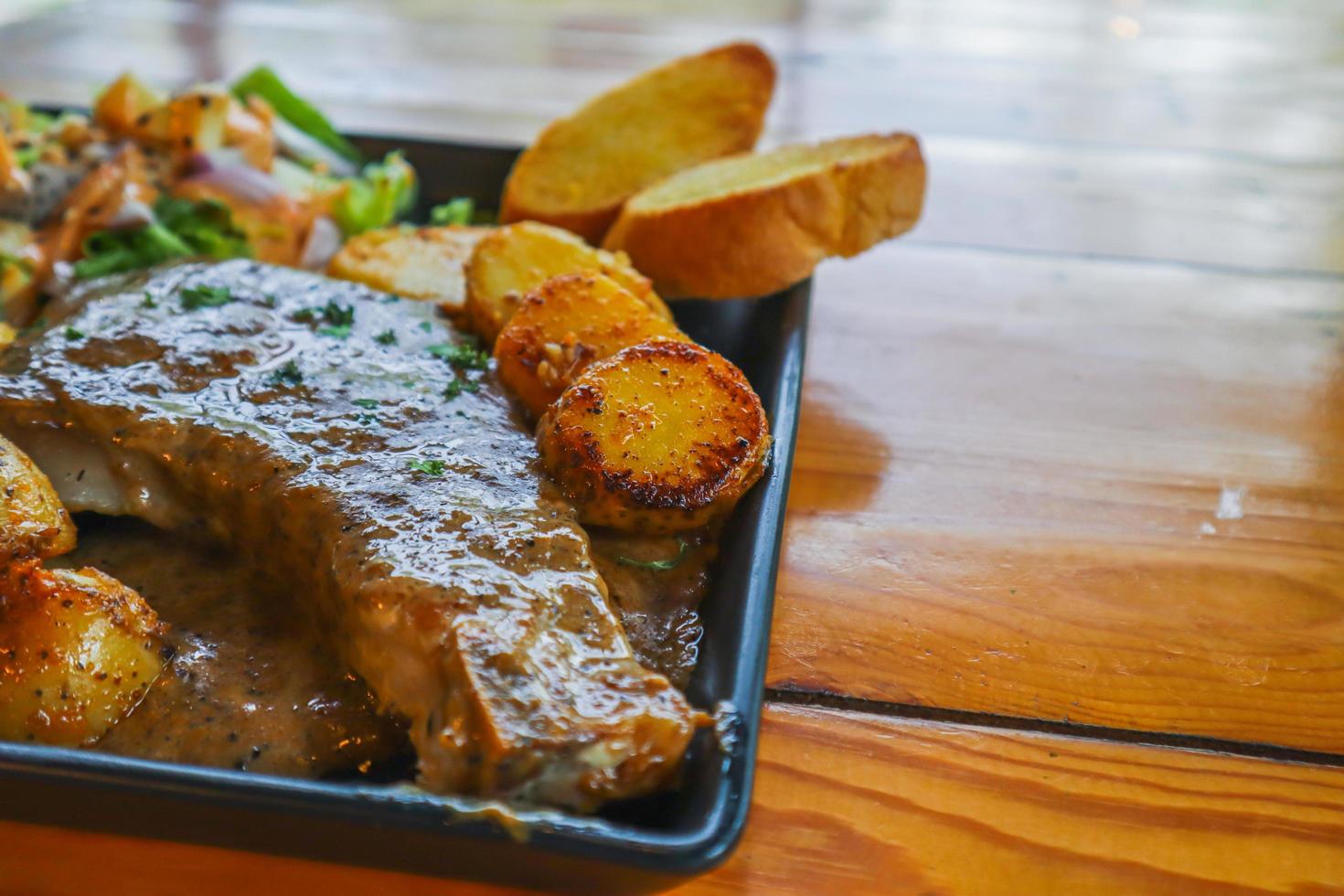 el bistec de ternera a la parrilla con salsa se sirve con una ensalada de frutas y verduras dentro de un plato de cerámica negra en la mesa del comedor para preparar el bistec para una cena festiva. foto