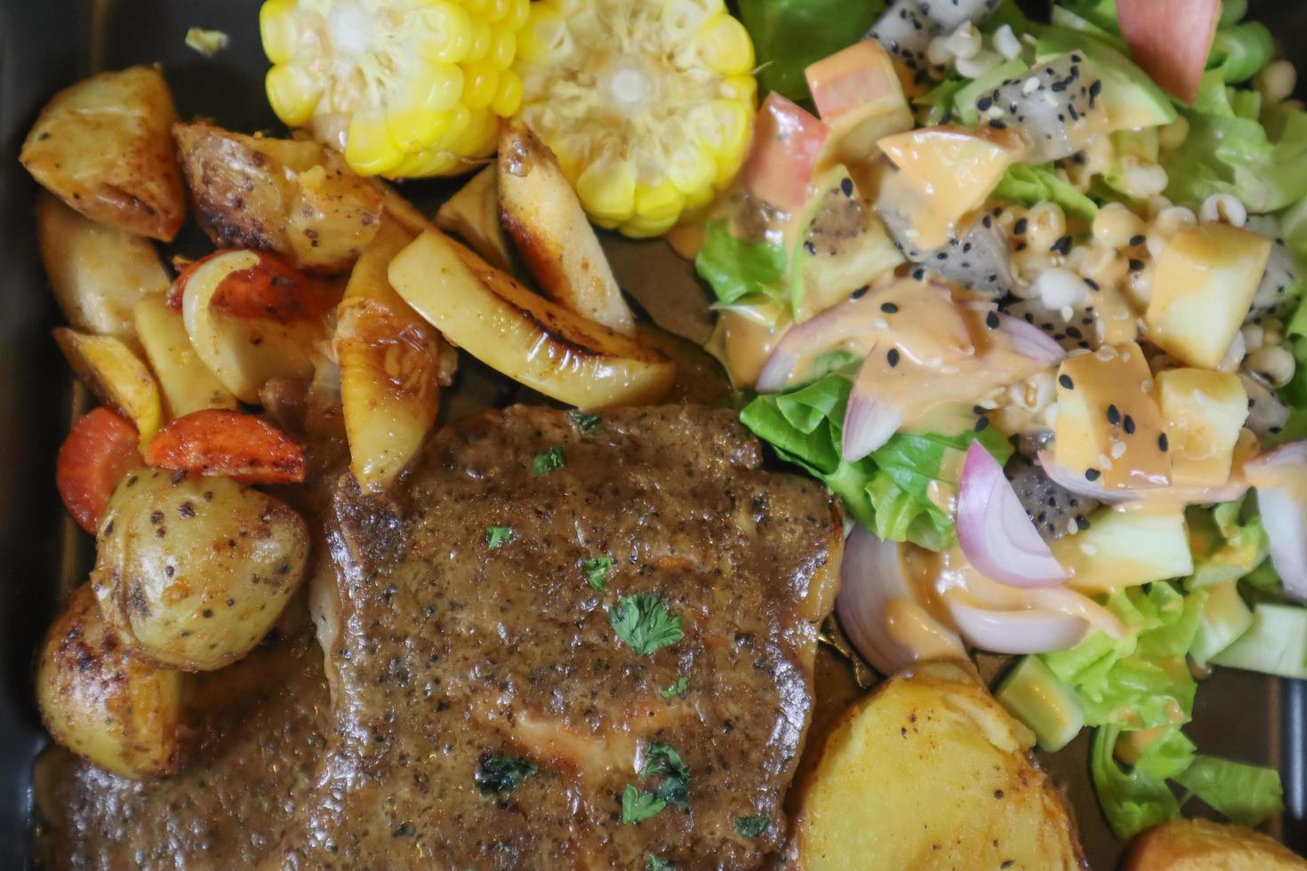 Grilled beef steak with gravy is served with a fruit and vegetable salad inside a black ceramic plate on the dining room table to prepare the steak for a festive dinner. photo