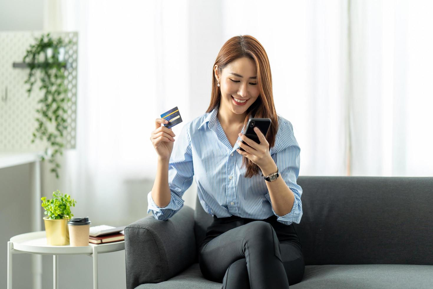 smiling asian woman holding smartphone and banking credit card, involved in online mobile shopping at home, happy female shopper purchasing goods or services in internet store. photo