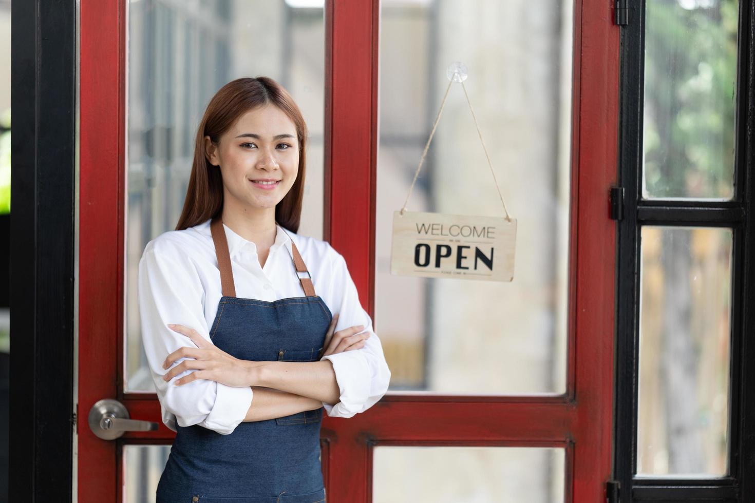 una mujer de negocios feliz asiática es una camarera en un delantal, el dueño del café se para en la puerta con un cartel abierto esperando a los clientes. concepto de pequeña empresa, cafeterías y restaurantes. foto