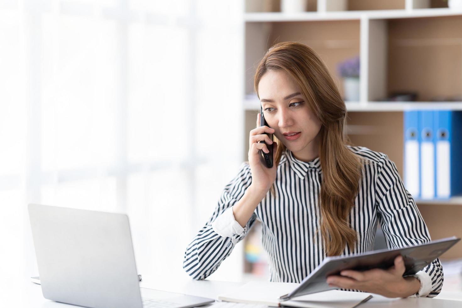 Young business woman on the phone at office. Business woman texting on the phone and working on laptop. Pretty young business woman sitting on workplace. Smiling business woman. photo