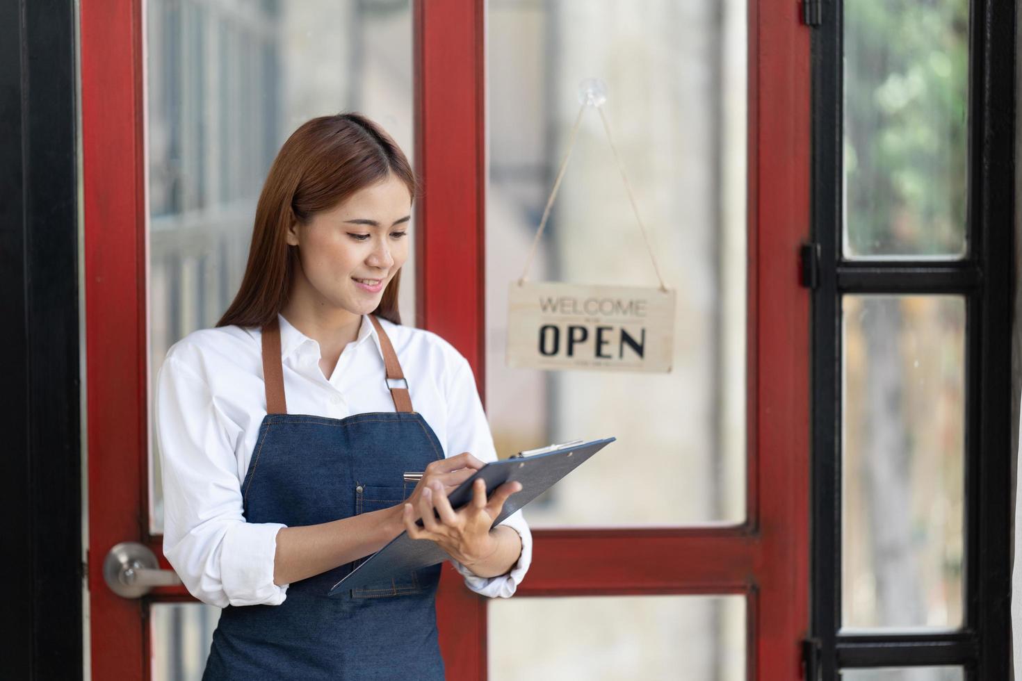 una mujer de negocios feliz asiática es una camarera en un delantal, el dueño del café se para en la puerta con un cartel abierto esperando a los clientes. concepto de pequeña empresa, cafeterías y restaurantes. foto