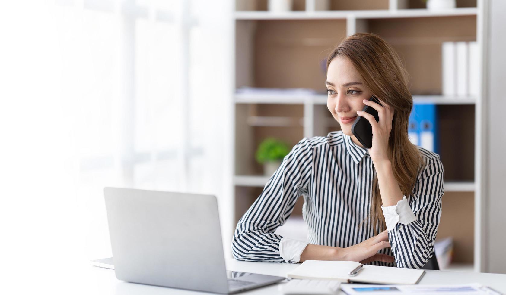 Young business woman on the phone at office. Business woman texting on the phone and working on laptop. Pretty young business woman sitting on workplace. Smiling business woman. photo
