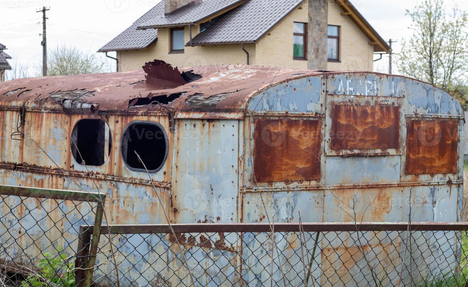 A dilapidated rotten and rusty metal hut on a work site. An old tin construction trailer with a round roof and a broken window from a meadow or pasture with a trailer drawbar. photo