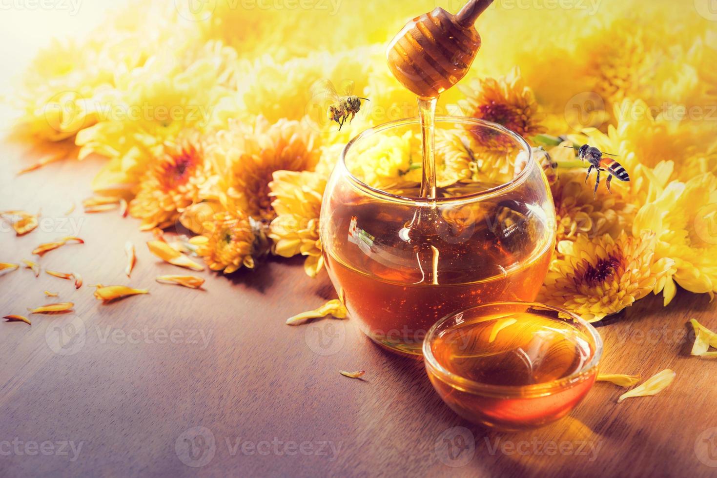 Honey in glass jar with bee flying and flowers on a wooden floor photo