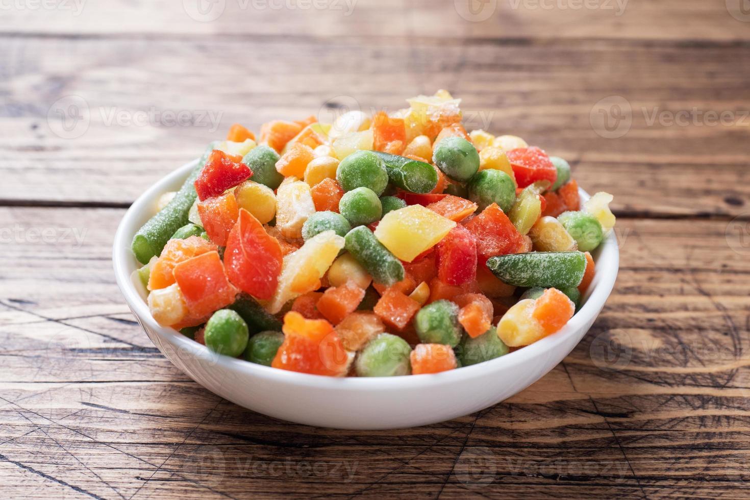 Chopped frozen vegetables in a plate on a wooden background. Corn peas pepper carrots. copy space. photo
