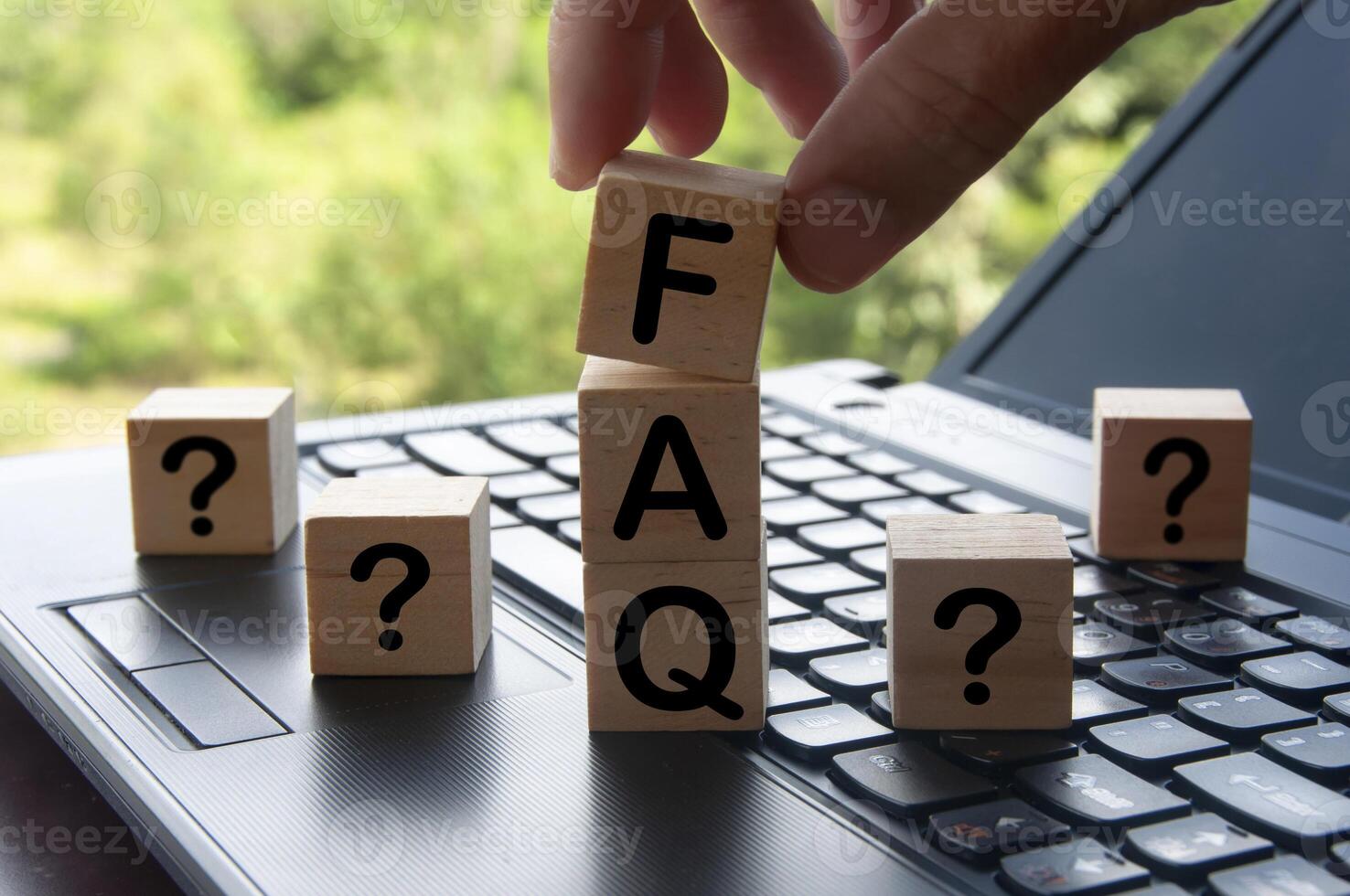 Hand holding FAQ word on wooden block on top of laptop with blurred nature background. Information concept photo