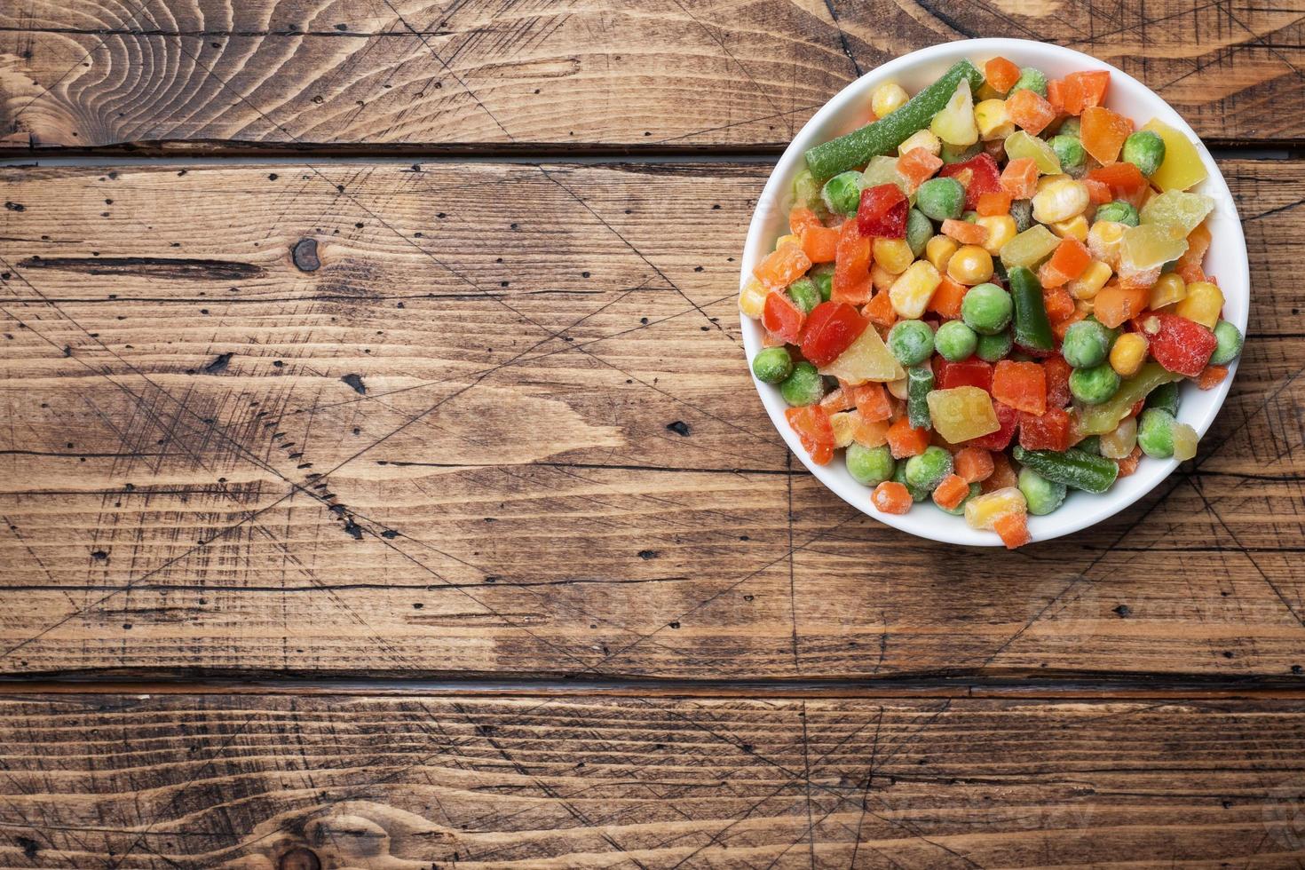 Chopped frozen vegetables in a plate on a wooden background. Corn peas pepper carrots. copy space. photo