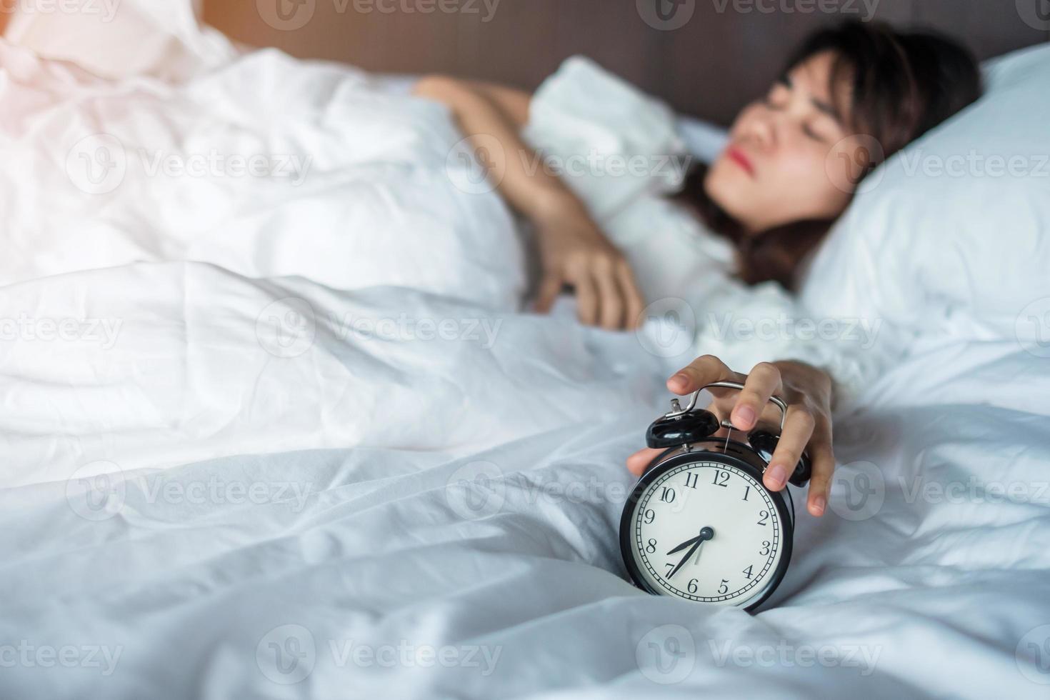 alarm clock and asian woman hand stop time in bed while sleeping, young adult female wake up late in the morning. Relaxing , sleepy, daily routine and have a nice day concepts photo