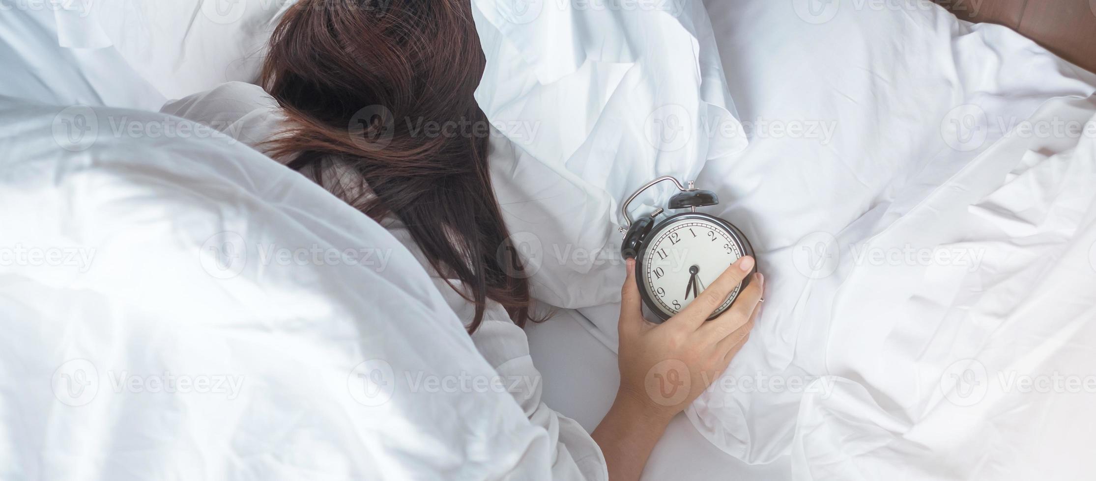 alarm clock and asian woman hand stop time in bed while sleeping, young adult female wake up late in the morning. Relaxing , sleepy, daily routine and have a nice day concepts photo