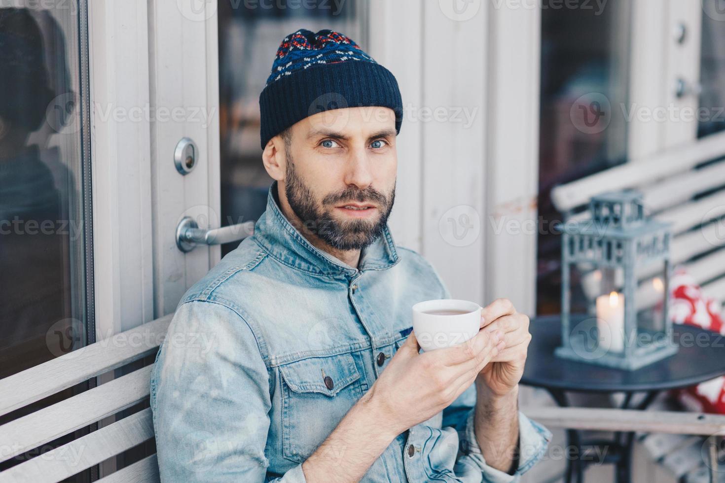 Close up shot of handsome serious bearded male, looks directly into camera with blue eyes and confident expression, drinks hot beverage, enjoys spare time. People, drinks, lifestyle concept. photo