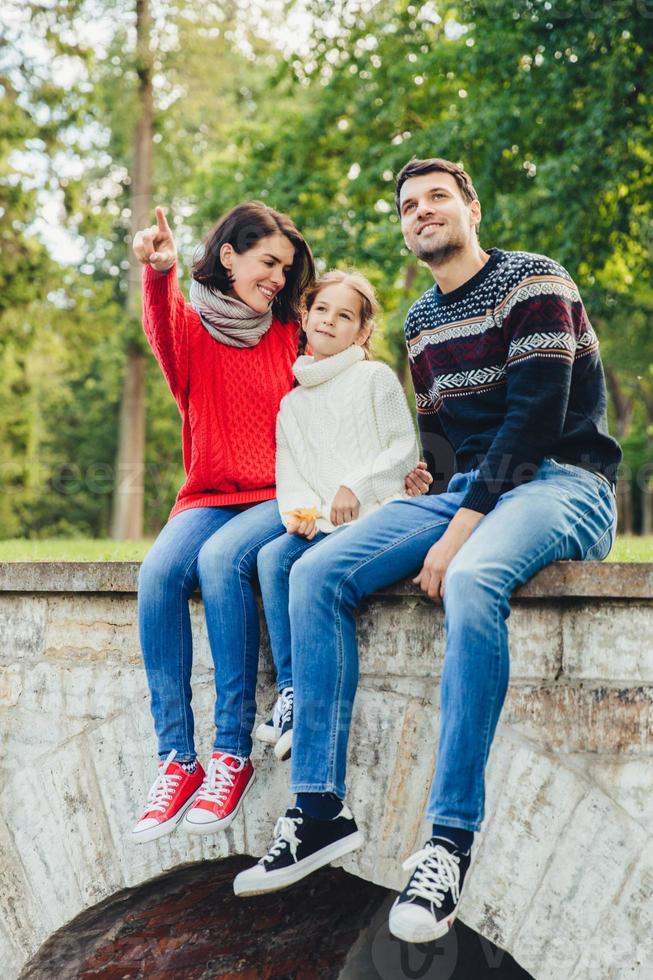 Carefree restful woman, man and small cute girl sits on bridge outdoors, admire sunrise. Affectionate mother shows to her little daughter beauty of nature. Family enjoys spending free time in park photo