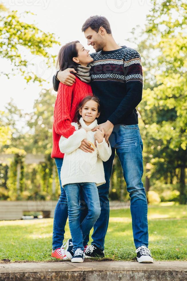 Young happy family of three stand together outdoors, embrace, have good relationships. Couple in love look at each other, hug their little daughter. Happiness and harmony in family life. photo