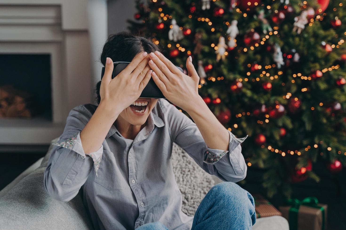 Joyful young woman in VR headset glasses sitting on cozy sofa next to decorated Xmas tree at home photo