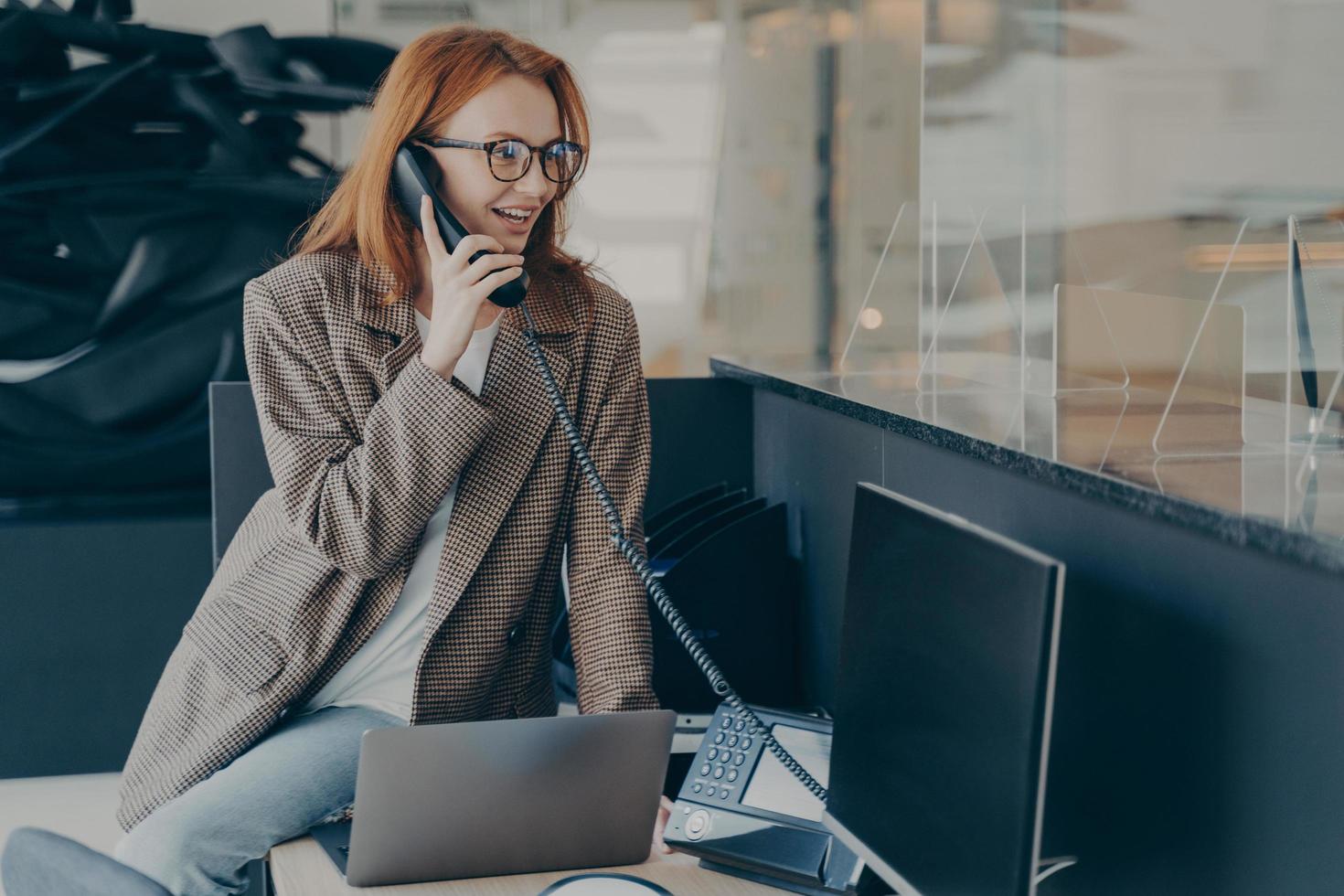 Woman in spectacles talking on phone while sitting at her workplace in office photo