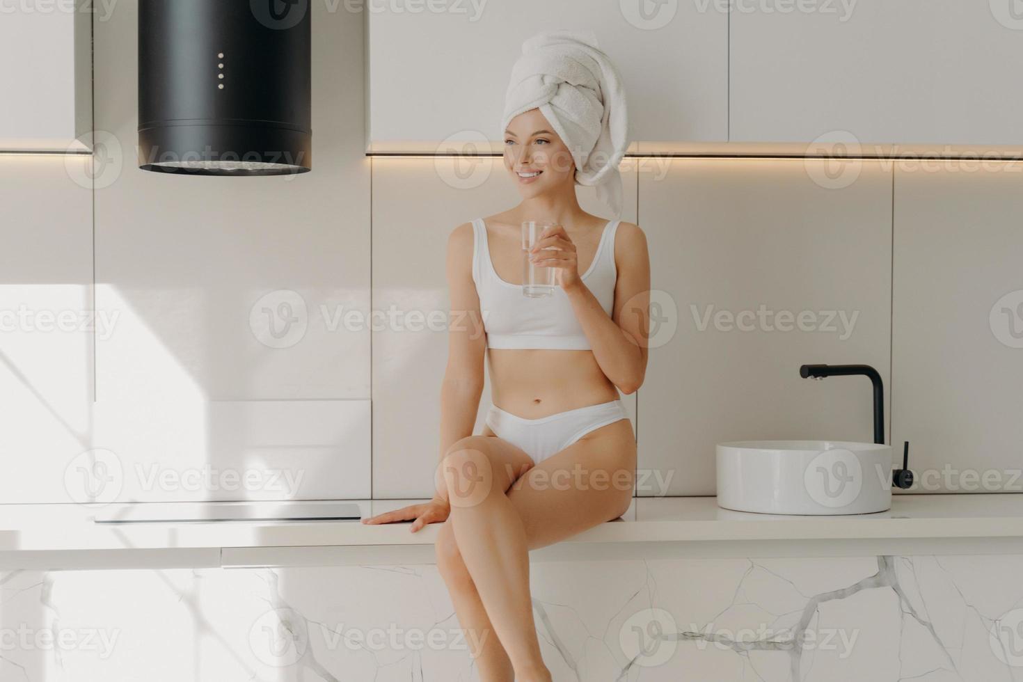 Slim sportive young girl sitting on white marble kitchen table top and drinking glass of mineral pure water photo